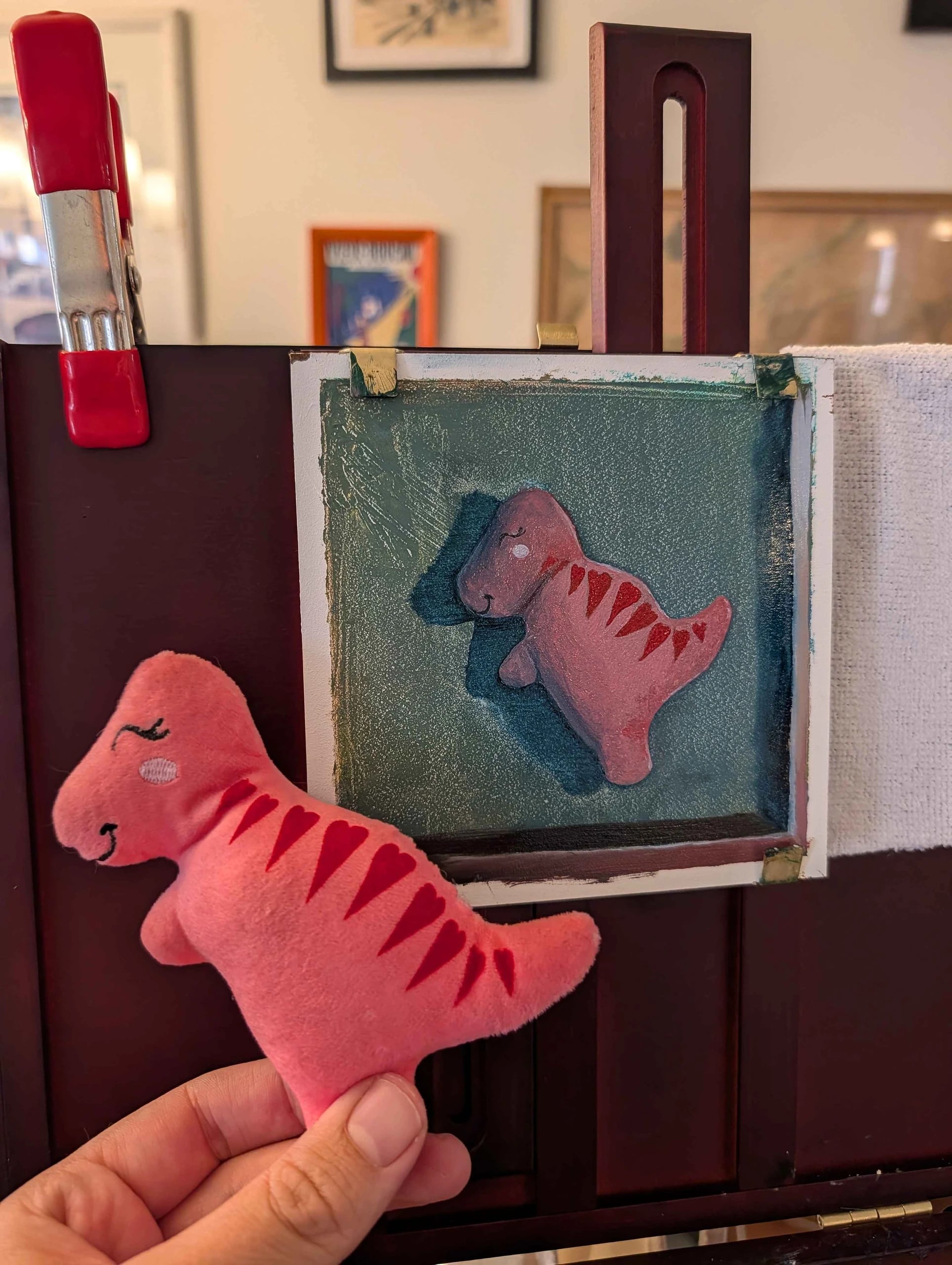 The pink dinosaur dog toy held up beside the finished painting on an easel for comparison.
