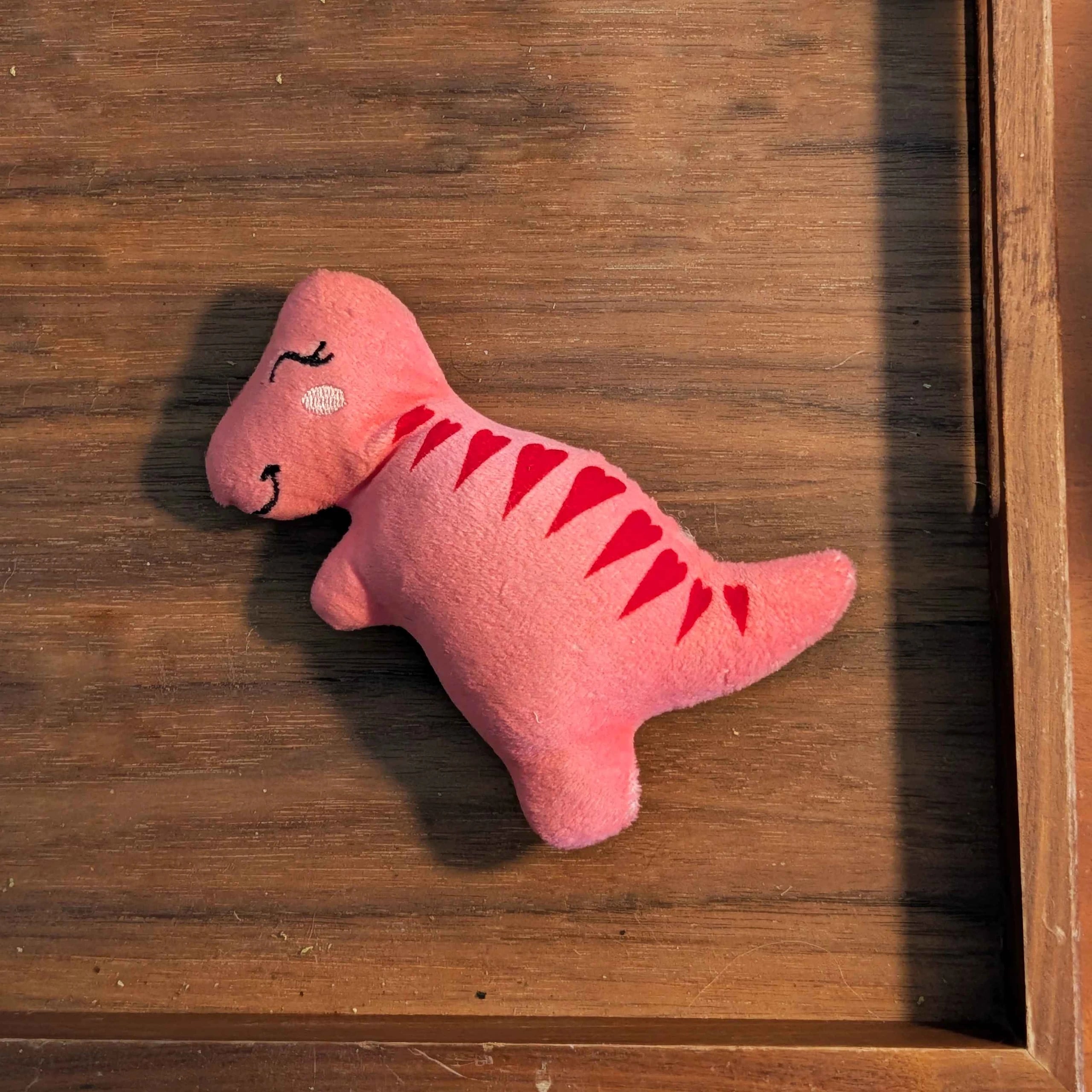 Reference photo: the pink plush dinosaur with red spikes lying in a wooden tray.
