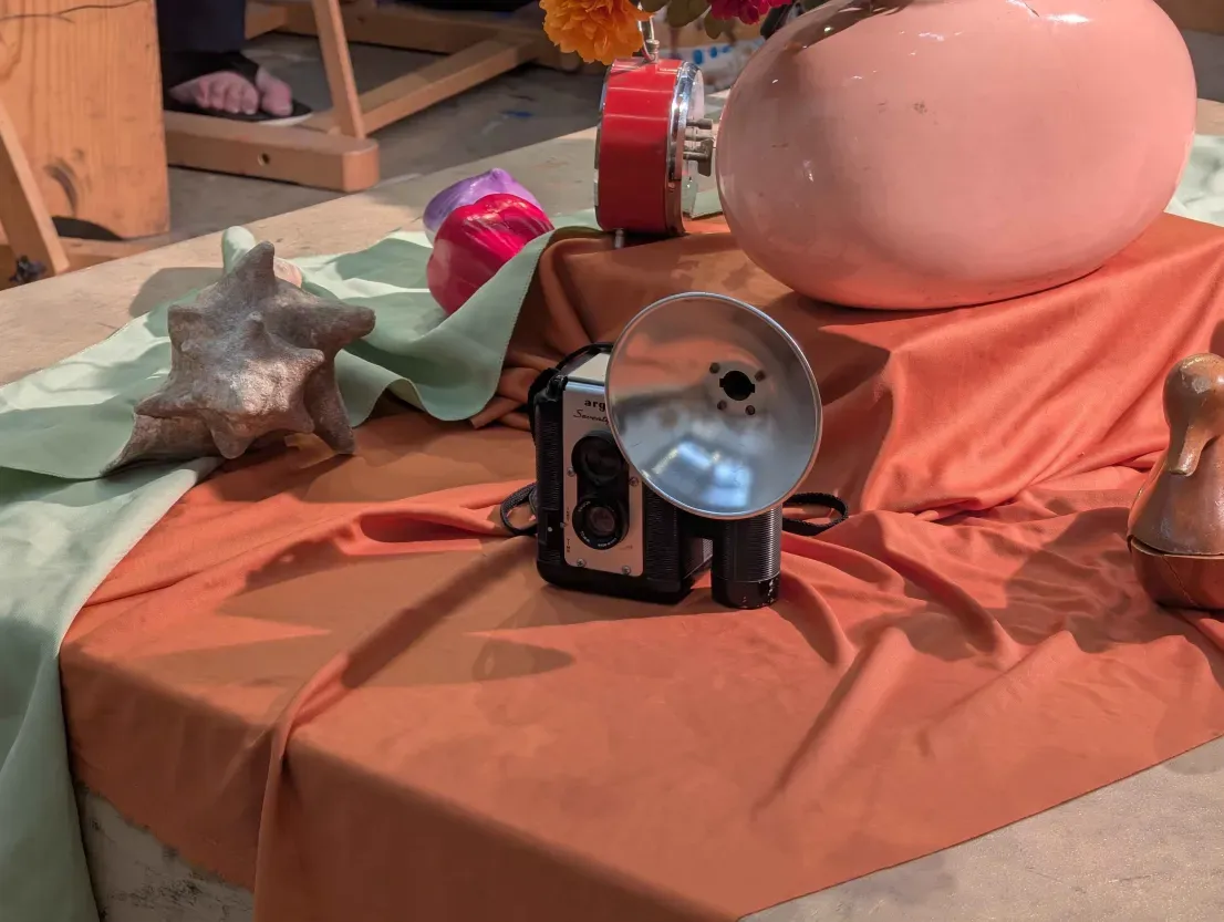 Reference photo of the class still life: a vintage flash camera on orange fabric beside a pink vase and a seashell.