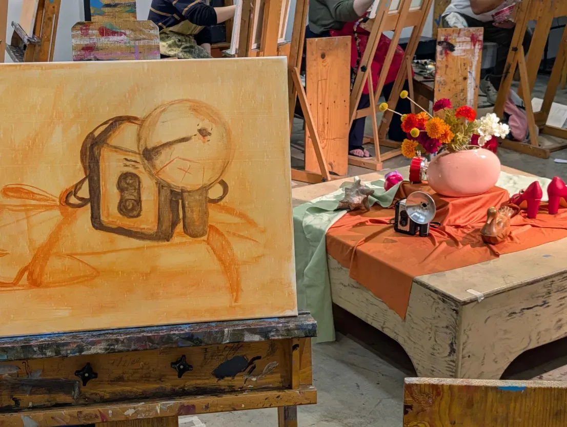 Photo of a painting studio: an easel holds an orange underpainting of a camera beside a still-life table with flowers and props.